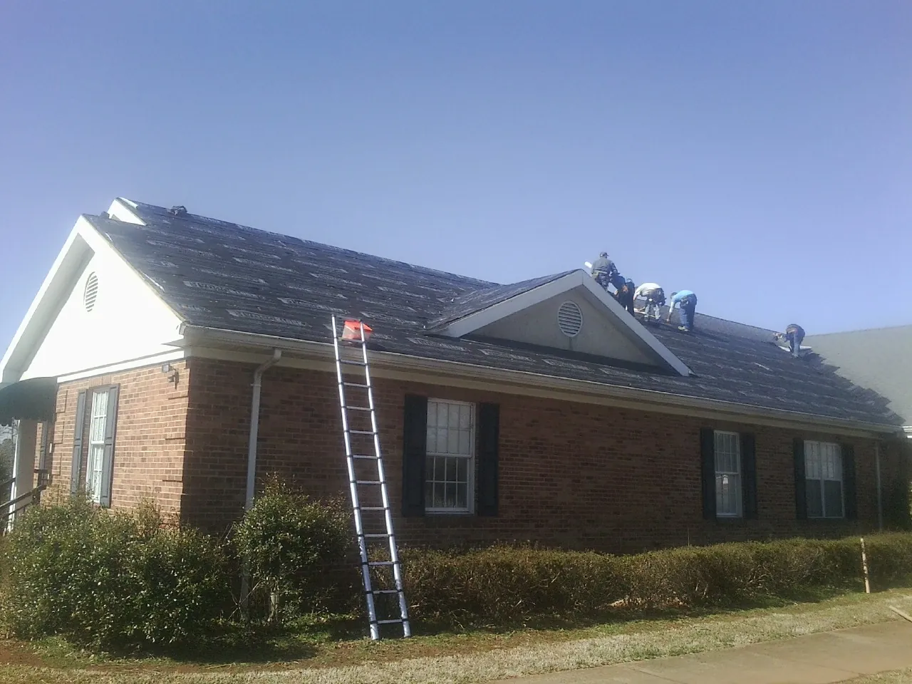 Crew tearing shingles off a brick ranch roof with a ladder set — Emergency Roof Repair work by our crew in Lillington