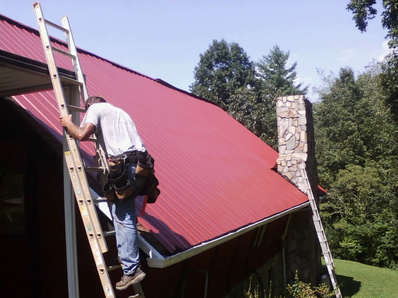 Roofer on a ladder beside a red metal roof and stone chimney — Soffit & Fascia Repair by Roofing Brothers in Lillington
